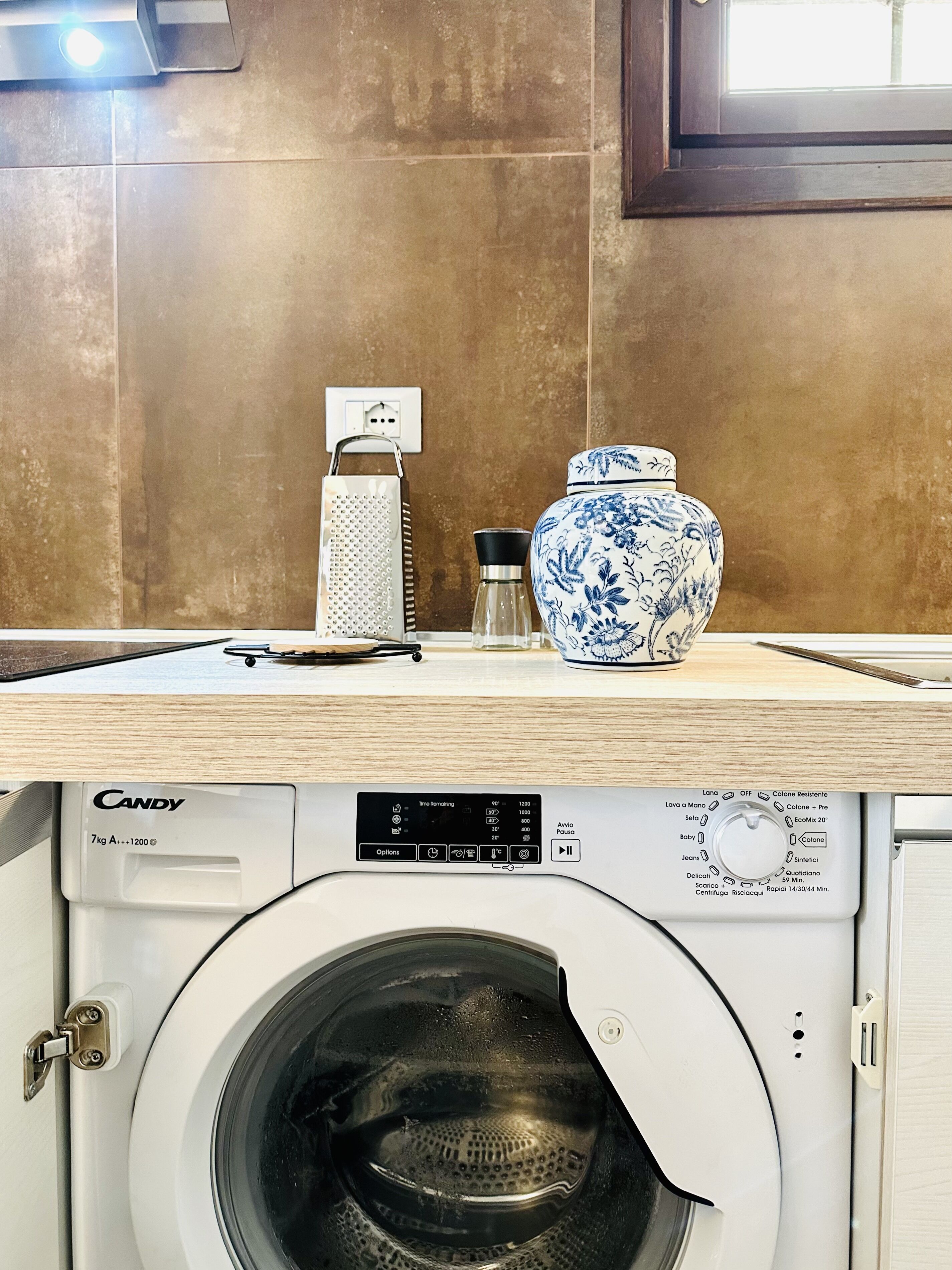 A kitchen with a washing machine, a grater, a salt shaker, and a blue and white vase on the counter.