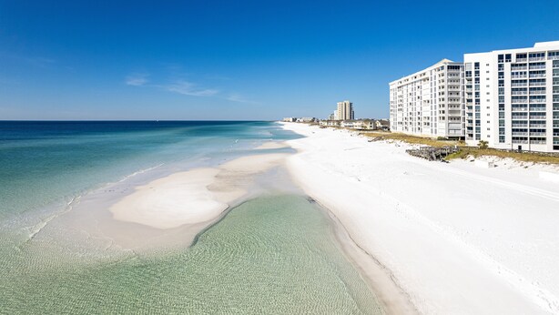 A beach with clear shallow waters, a sandy shore, and a row of white buildings with multiple floors and balconies. - Shoreline Towers 1052 - Beautiful 2 BR with Gulf views (Destin)