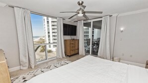 A bedroom with a large bed, a ceiling fan, a wooden dresser, and a view of a beach and high-rise buildings through a sliding glass door. - Shoreline Towers 1052 - Beautiful 2 BR with Gulf views (Destin)