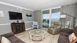 A living room with a TV, a bookshelf, a white chair, a leather armchair, and a glass coffee table. - Shoreline Towers 1084 - Beautiful 2 BR with Gulf views and on the beach (Destin)
