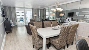 A modern hotel room with a dining area featuring a white table and woven chairs, a large window with curtains, and a view of the cityscape. - Shoreline Towers 1084 - Beautiful 2 BR with Gulf views and on the beach (Destin)