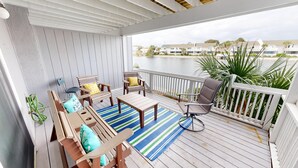 Une terrasse extérieure couverte, aménagée avec des meubles en bois, un tapis à rayures et offrant une vue sur une étendue d’eau et des bâtiments en arrière-plan.