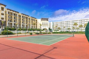 A tennis court surrounded by a hotel building with multiple balconies and palm trees.