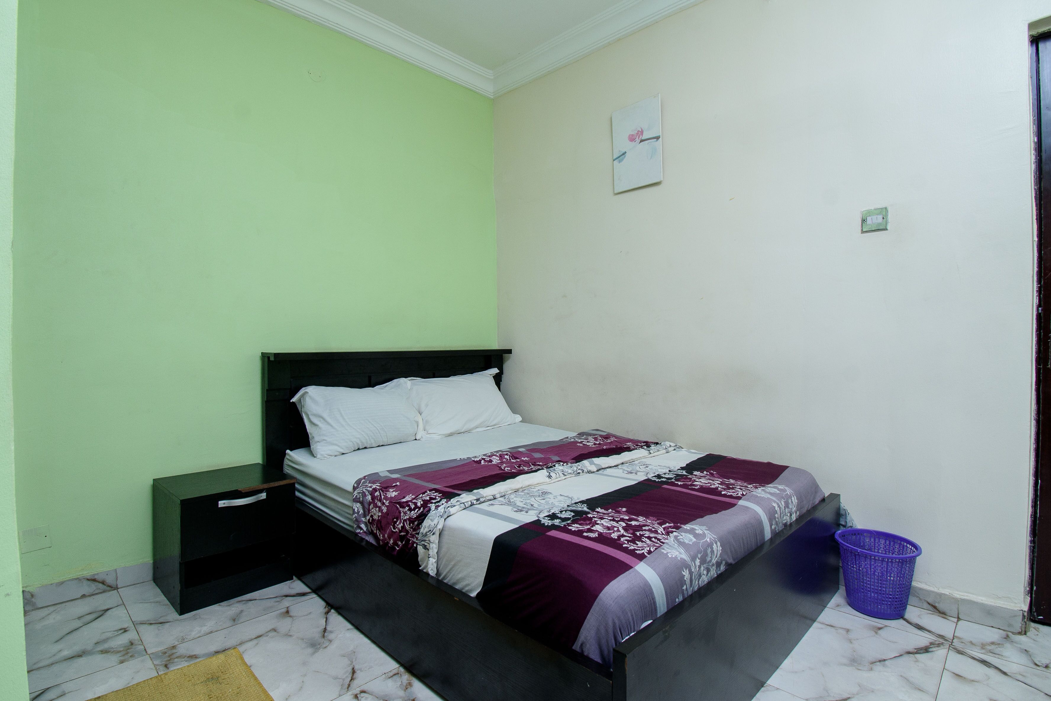 A bedroom with a bed, a nightstand, a wall-mounted light switch, and a door.