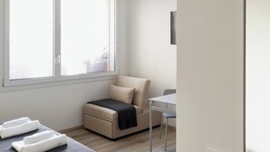 Une petite pièce lumineuse, avec un canapé, une table et une fenêtre équipée de stores.
