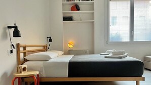 Un lit simple avec un cadre en bois, une table de chevet avec une lampe, un petit tabouret et une étagère contenant divers objets.