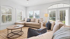 A living room with a beige sectional sofa, a round coffee table, and a lamp. There are large windows with white blinds and a view of greenery outside.