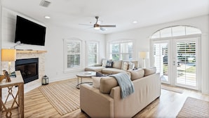 A spacious living room with a fireplace, a large sofa, a television, and large windows with shutters.