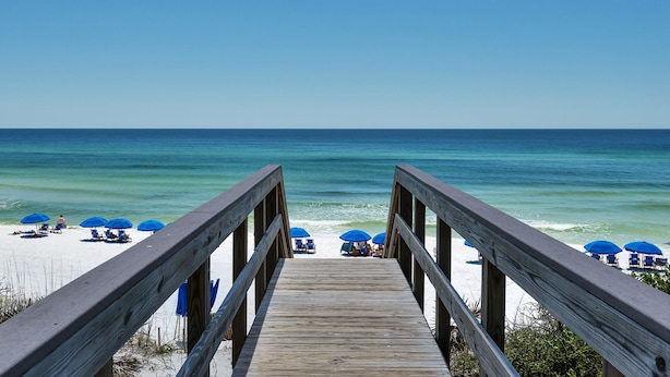 A wooden boardwalk leading to a beach with blue umbrellas and lounge chairs.