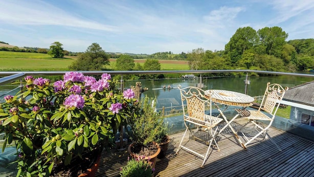 A deck with a glass balustrade overlooking a lake, surrounded by greenery and flowers. - The Boathouse (Bath)
