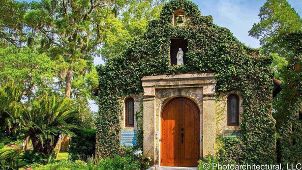 Una pequeña iglesia con cruz, campana y estatua, rodeada de abundante vegetación.