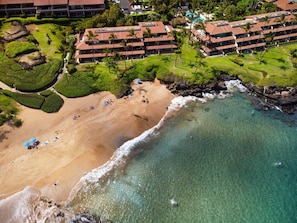 Vue aérienne d’un hôtel situé sur la côte, avec une plage de sable, des eaux turquoise et limpides, et une végétation luxuriante.