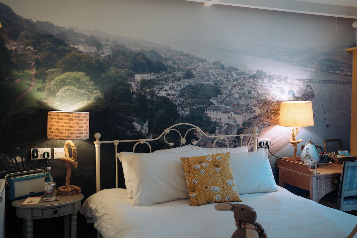 A hotel room with a bed, two bedside tables, a lamp, a map on the wall, and a view of a town.