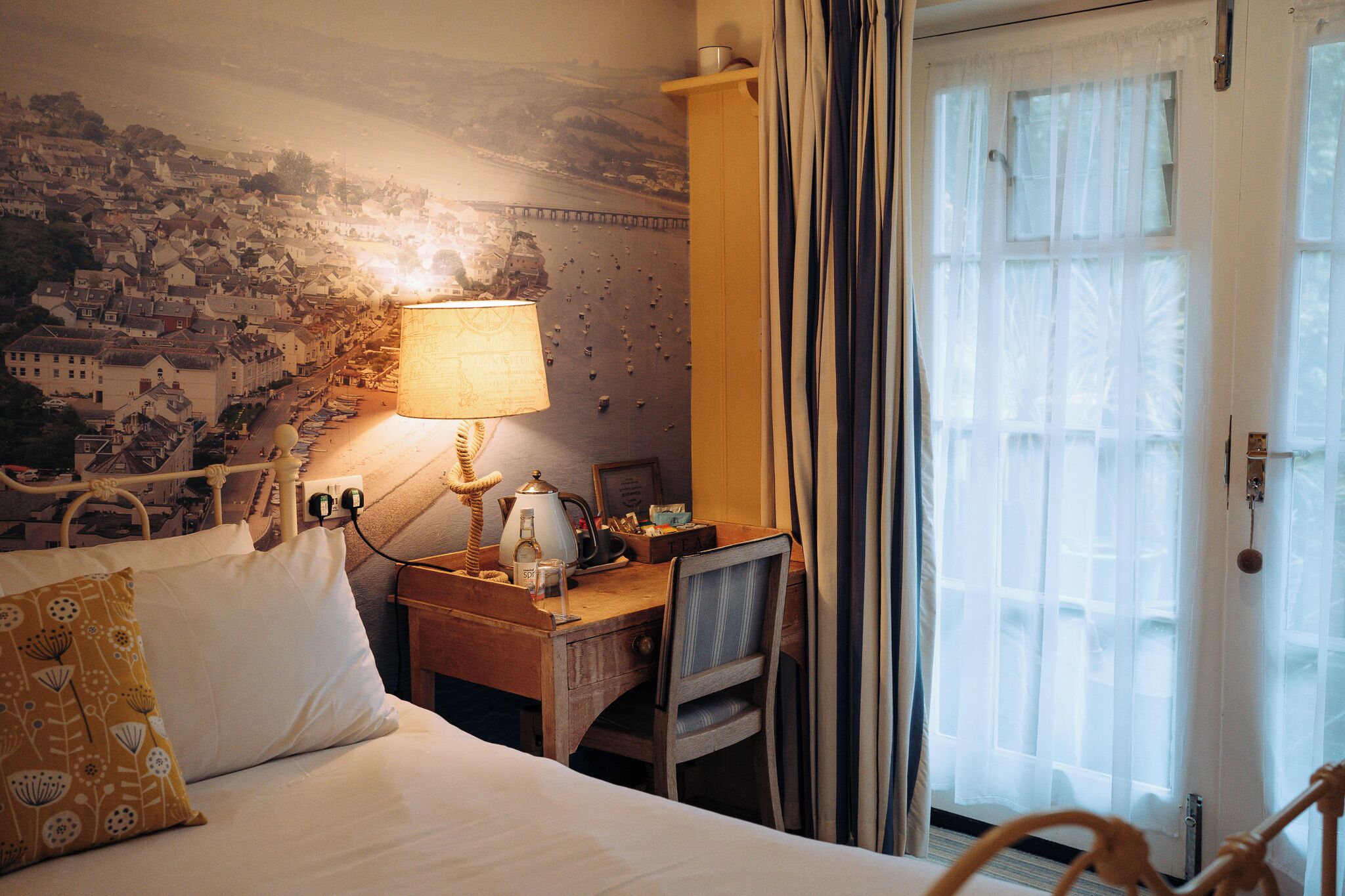 A hotel room with a bed, a desk with a lamp, a chair, and a view of a coastal town through a window.