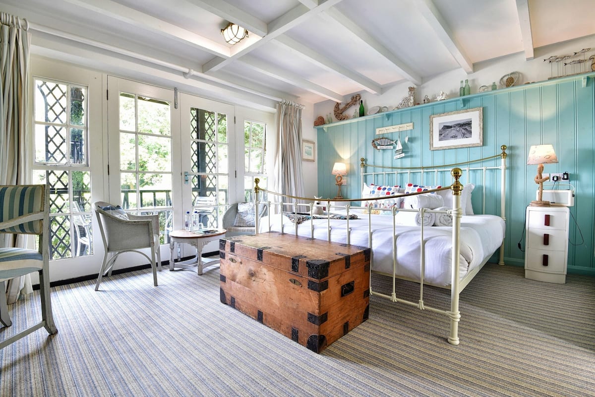 A bedroom with a four-poster bed, a wooden chest, and a view of the outdoors through large windows.