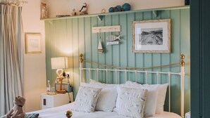 A neatly made bed with white linens and a metal frame, a bedside table with a lamp, and a wall-mounted shelf with decorative items. - The Ness House (Teignmouth)