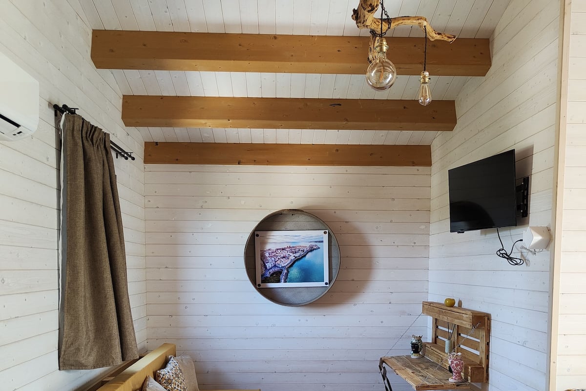 Tunnelmallinen huone, jossa on puinen katto, seinälle asennettu pyöreä televisio, tyynyillä varustettu sohva, pieni puinen pöytä ja tuoli.