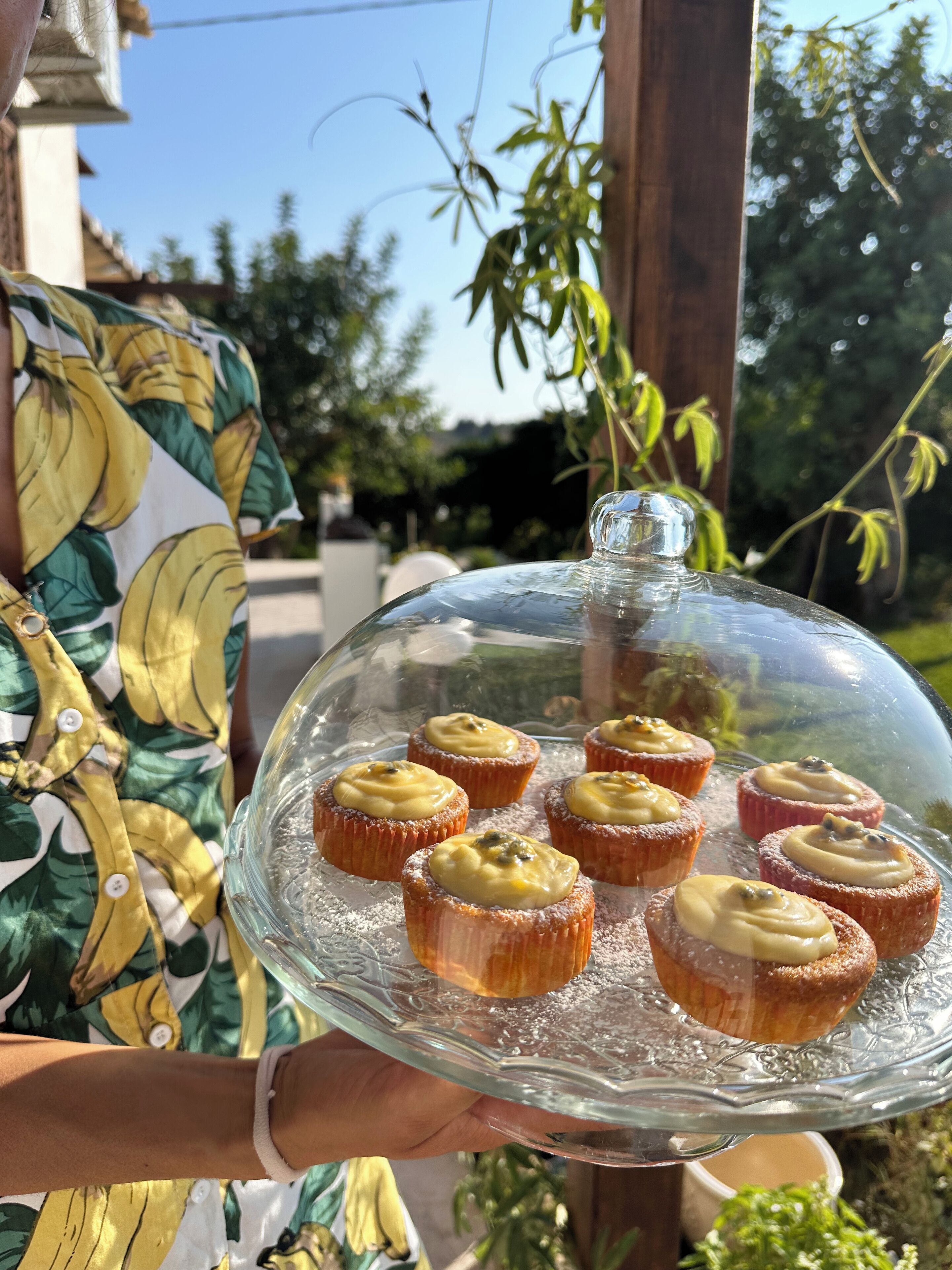 A person holding a glass dome display case with cupcakes.
