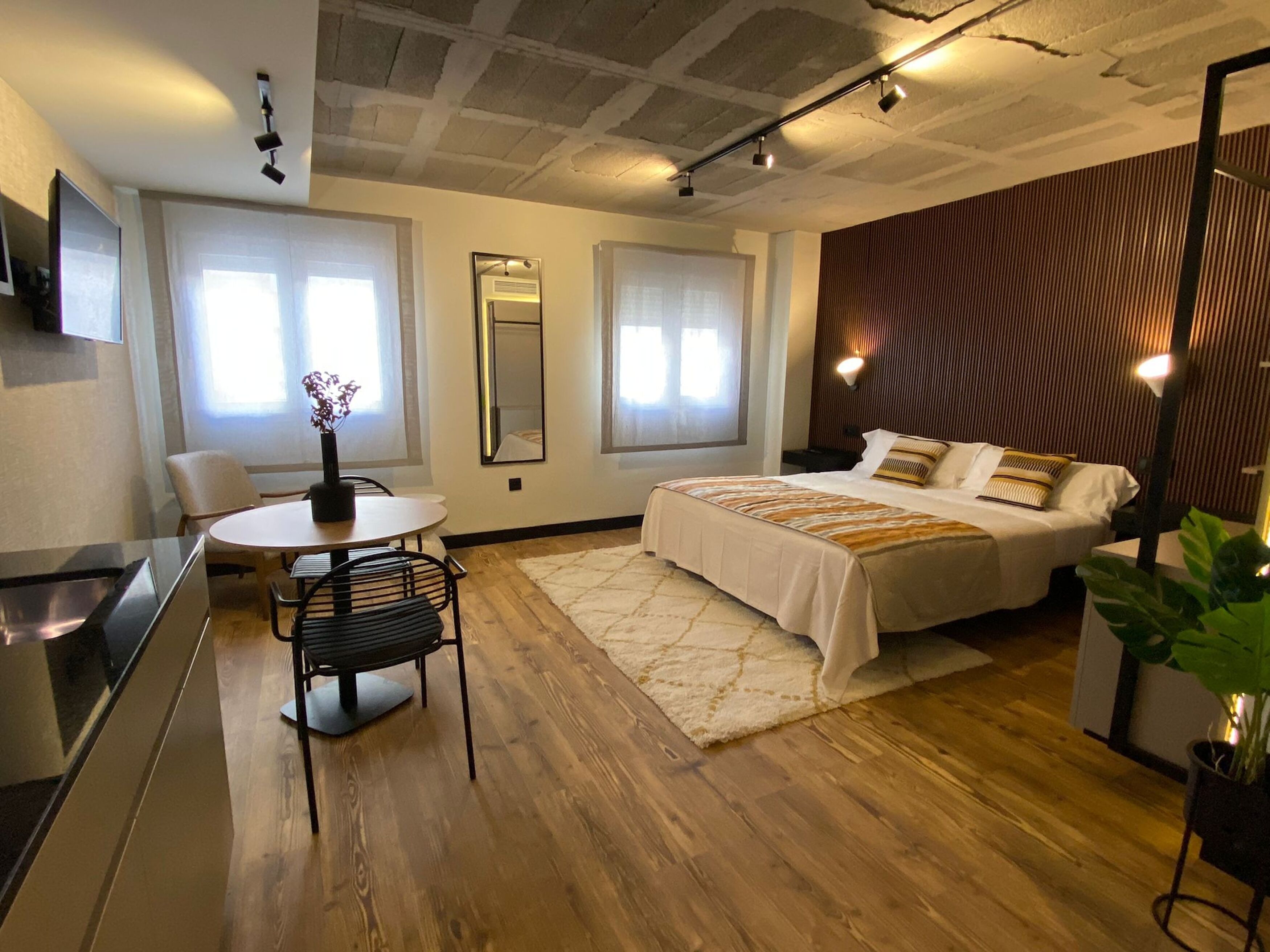 A modern hotel room with a bed, a desk, and a chair. There is a large mirror on the wall and a window with curtains.
