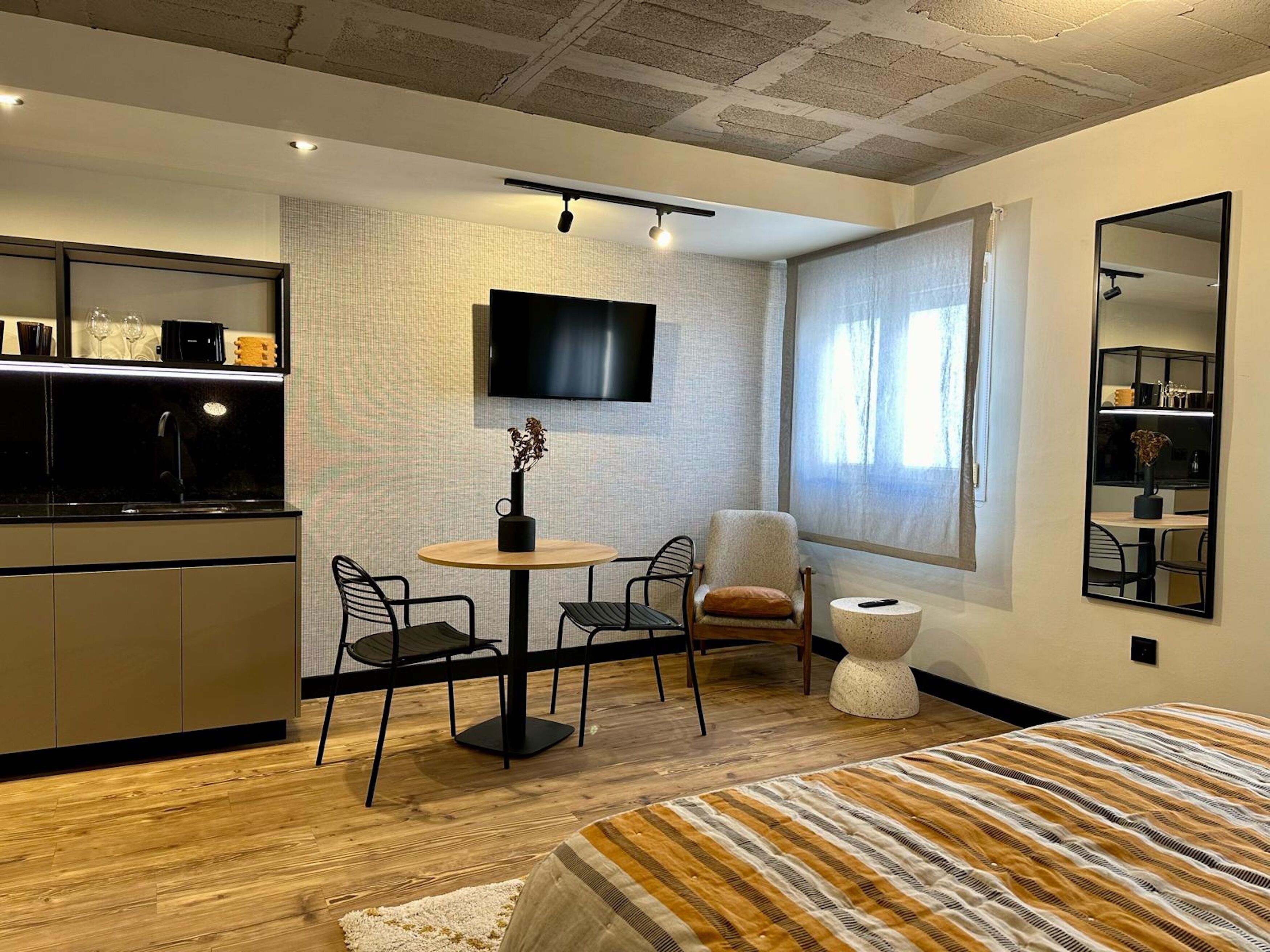 Una habitación de hotel moderna con suelo de madera, una cama con ropa de cama a rayas, una mesa redonda con sillas, un televisor montado en la pared y un amplio ventanal con cortinas.