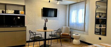 Una habitación de hotel moderna con suelo de madera, una cama con ropa de cama a rayas, una mesa redonda con sillas, un televisor montado en la pared y un amplio ventanal con cortinas.