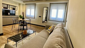 A modern living room with a sofa, a dining table, and chairs. There is a kitchen area in the background with a countertop and cabinets. - BUBU Apartments By Exclusive Homes (Málaga)