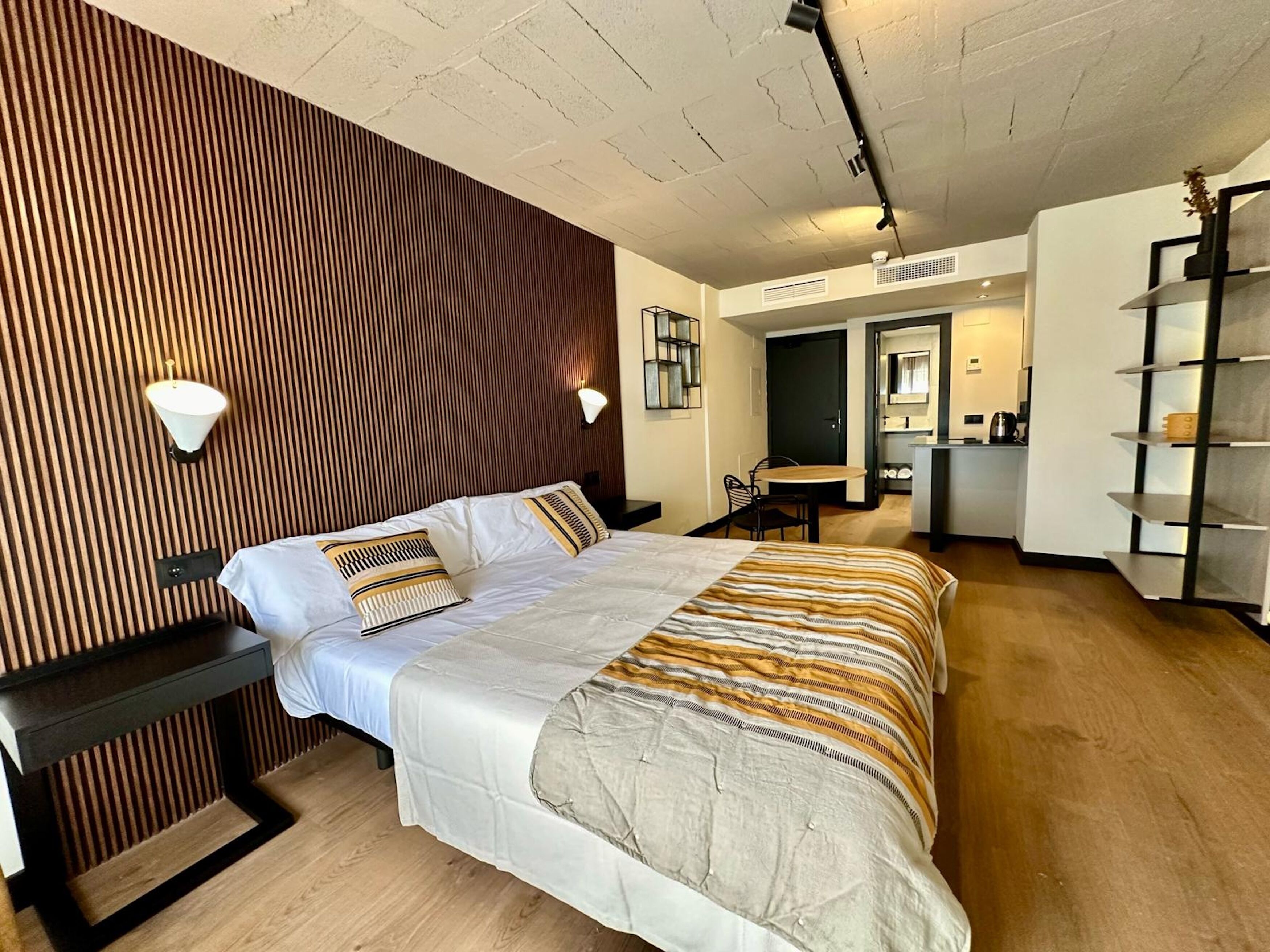 Una habitación de hotel moderna con una cama grande, un panel de madera decorativo, un escritorio y una kitchenette.