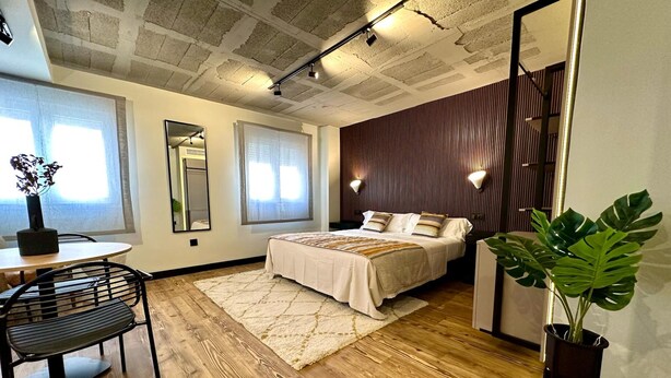 A modern bedroom with a wooden floor, a bed with white bedding, a round dining table with chairs, a large mirror, and a potted plant. - BUBU Apartments By Exclusive Homes (Málaga)