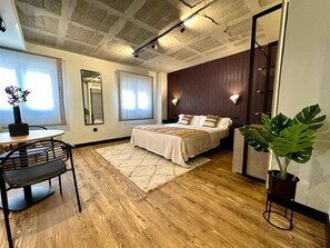 Un dormitorio moderno con suelo de madera, una cama con ropa de cama blanca, una mesa de comedor redonda con sillas, un espejo grande y una planta en maceta.
