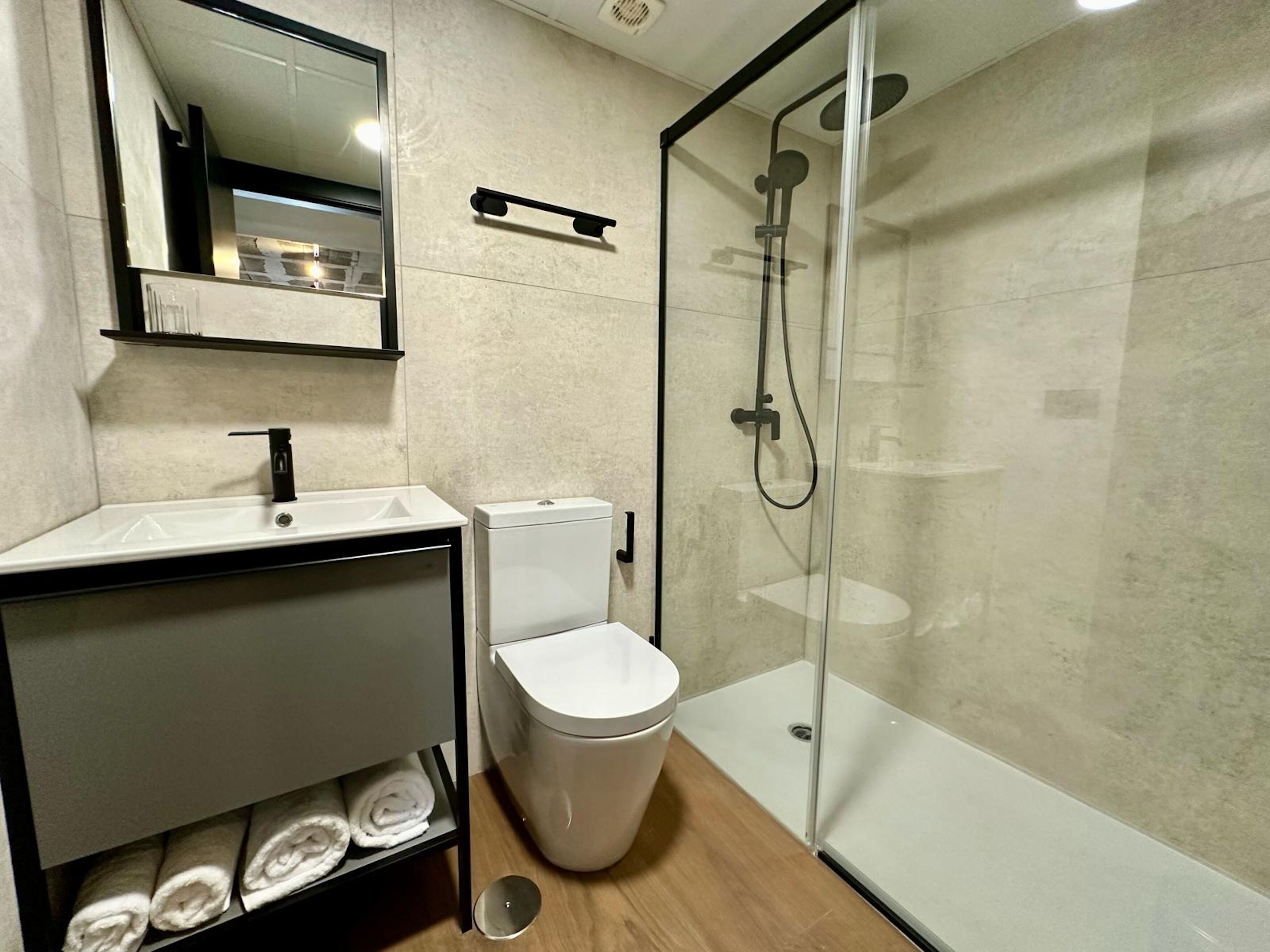 Une salle de bain moderne avec une douche à parois de verre, des toilettes blanches, un lavabo avec un miroir au-dessus et une étagère où sont pliés des serviettes.