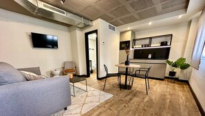 A modern living room with a grey sofa, a coffee table, and a dining area with a small table and chairs. There is a kitchen area with a microwave and a sink. - BUBU Apartments By Exclusive Homes (Málaga)