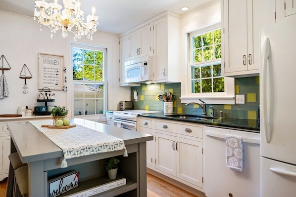 A modern kitchen with white cabinets, a central island, a chandelier, and a view of greenery outside.