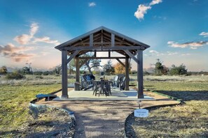 A covered outdoor seating area with tables and chairs, a barbecue grill, and a welcome sign. - Holy Cow Cottage w/Amazing Hill Country Views (Fredericksburg)