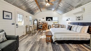 A spacious room with a high ceiling, a bed, a sofa, a desk, and a kitchen area. - Holy Cow Cottage w/Amazing Hill Country Views (Fredericksburg)