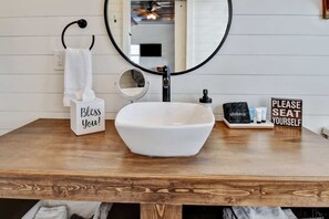 A bathroom with a wooden vanity, a white sink, a round mirror, a towel rack, a tissue box, a makeup station, and a sign that says "PLEASE SEAT YOURSELF". - Holy Cow Cottage w/Amazing Hill Country Views (Fredericksburg)
