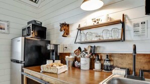 A kitchen with a wooden countertop, a stainless steel refrigerator, a microwave, and a sink. There are utensils in a container, a jar of utensils, and a shelf with glassware. - Holy Cow Cottage w/Amazing Hill Country Views (Fredericksburg)