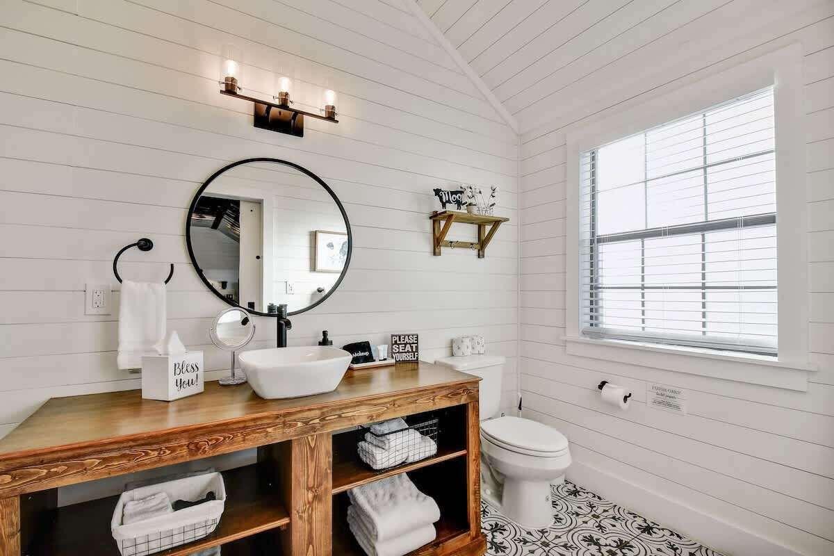 Une salle de bain avec un meuble-lavabo en bois, un miroir rond, un lavabo blanc et des toilettes. Il y a une fenêtre avec des stores et une étagère sur laquelle sont posés des objets décoratifs.