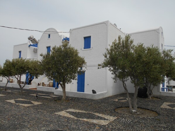 A white building with blue doors and windows, surrounded by trees and a cobblestone ground. - The Three Anchors Rooms (Santorini)