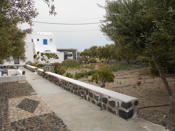 A white building with a blue door, surrounded by a stone wall and a garden with various plants. - The Three Anchors Rooms (Santorini)