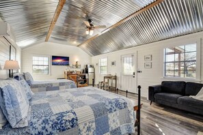 Een ruime kamer met een hoge plafond, een bed met een blauw-witte quilt, een donkere bank en een keuken met een koelkast en fornuis.