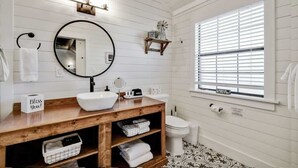 Une salle de bain avec un meuble-lavabo en bois, un miroir rond, un lavabo blanc et une fenêtre avec des stores.