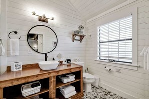 Un baño con tocador de madera, espejo redondo, lavabo blanco y ventana con persianas.