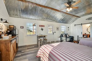 A rustic room with a bed, a desk, a chair, a small table, and a kitchen area.
