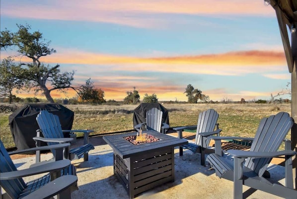 A patio area with Adirondack chairs, a fire pit, and a grill, set against a sunset backdrop.