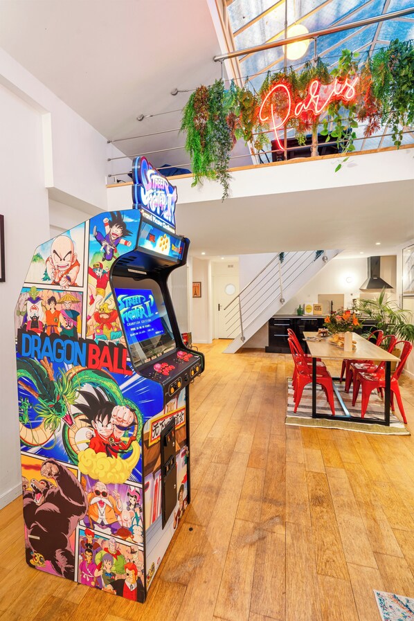 A gaming arcade with Dragon Ball characters, a neon Paris sign, and a staircase leading to a balcony with plants.