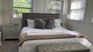 Een keurig opgemaakt bed met een gestoffeerde bank, twee nachtkastjes met lampen, en ramen met zonwering.