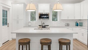 Una cocina moderna con armarios blancos, una isla central con taburetes, iluminación colgante y una campana extractora de acero inoxidable.