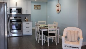 A kitchen with white cabinets, a stainless steel refrigerator, a microwave, and an oven. There is a dining table with white chairs and a wicker chair. - Sunset Point Marina - Dolphin (Manasota Key)