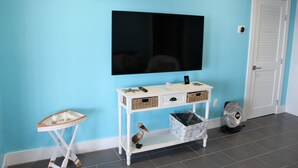 Una stanza con una parete blu, un tavolino consolle bianco con cesti, un piccolo tavolo bianco, una televisione e una porta con un cartello.