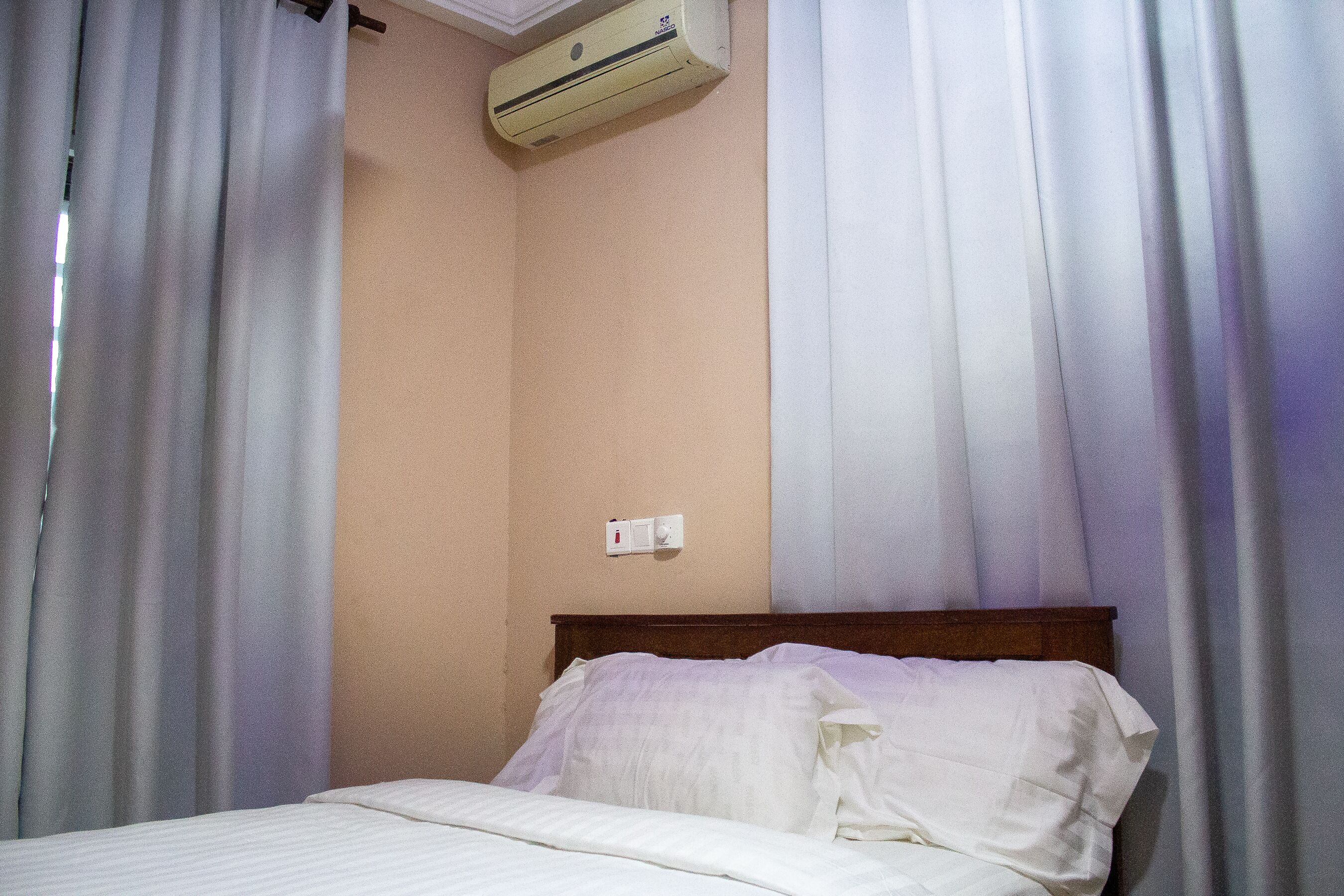 A hotel room with a bed, curtains, an air conditioning unit, and a wall switch.